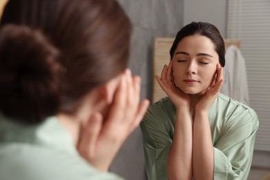 Photo of Young woman massaging her face near mirror in bathroom