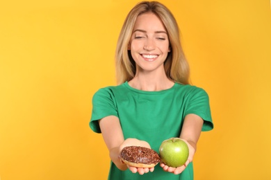 Photo of Woman choosing between doughnut and healthy apple on yellow background