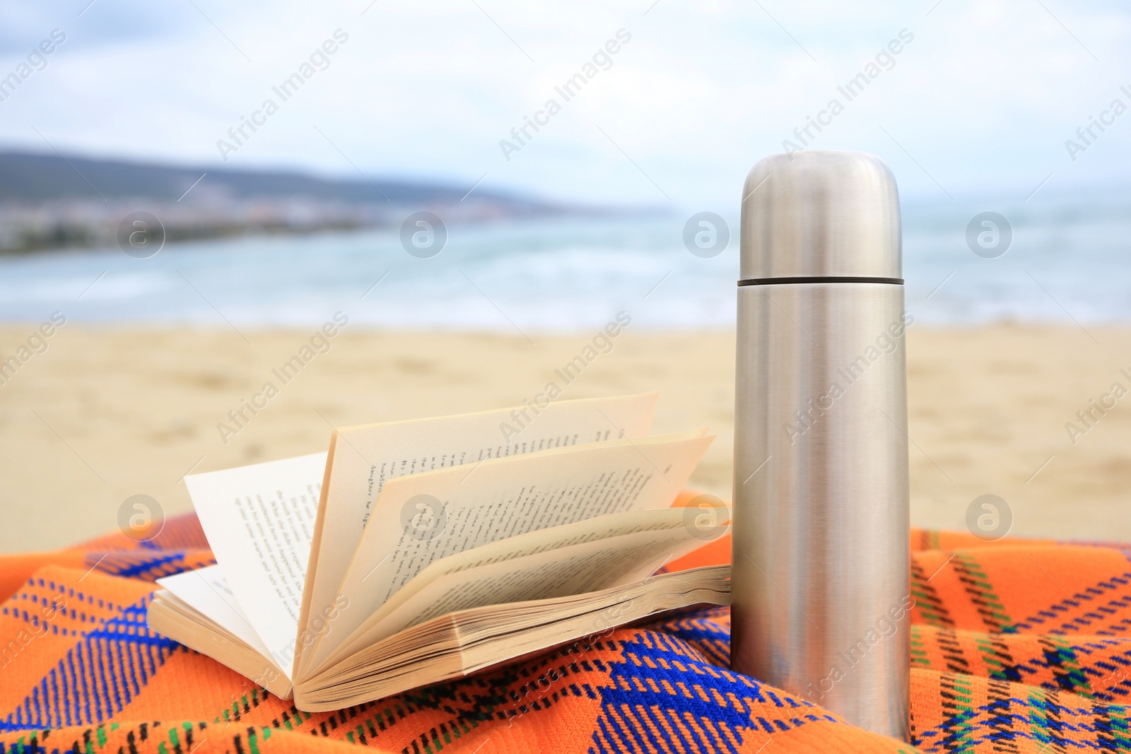 Photo of Metallic thermos with hot drink, open book and plaid on sandy beach near sea