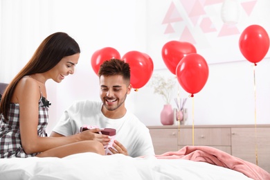 Photo of Young man presenting gift to his girlfriend in bedroom decorated with air balloons. Celebration of Saint Valentine's Day