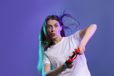 Surprised woman playing video games with controller on violet background, space for text