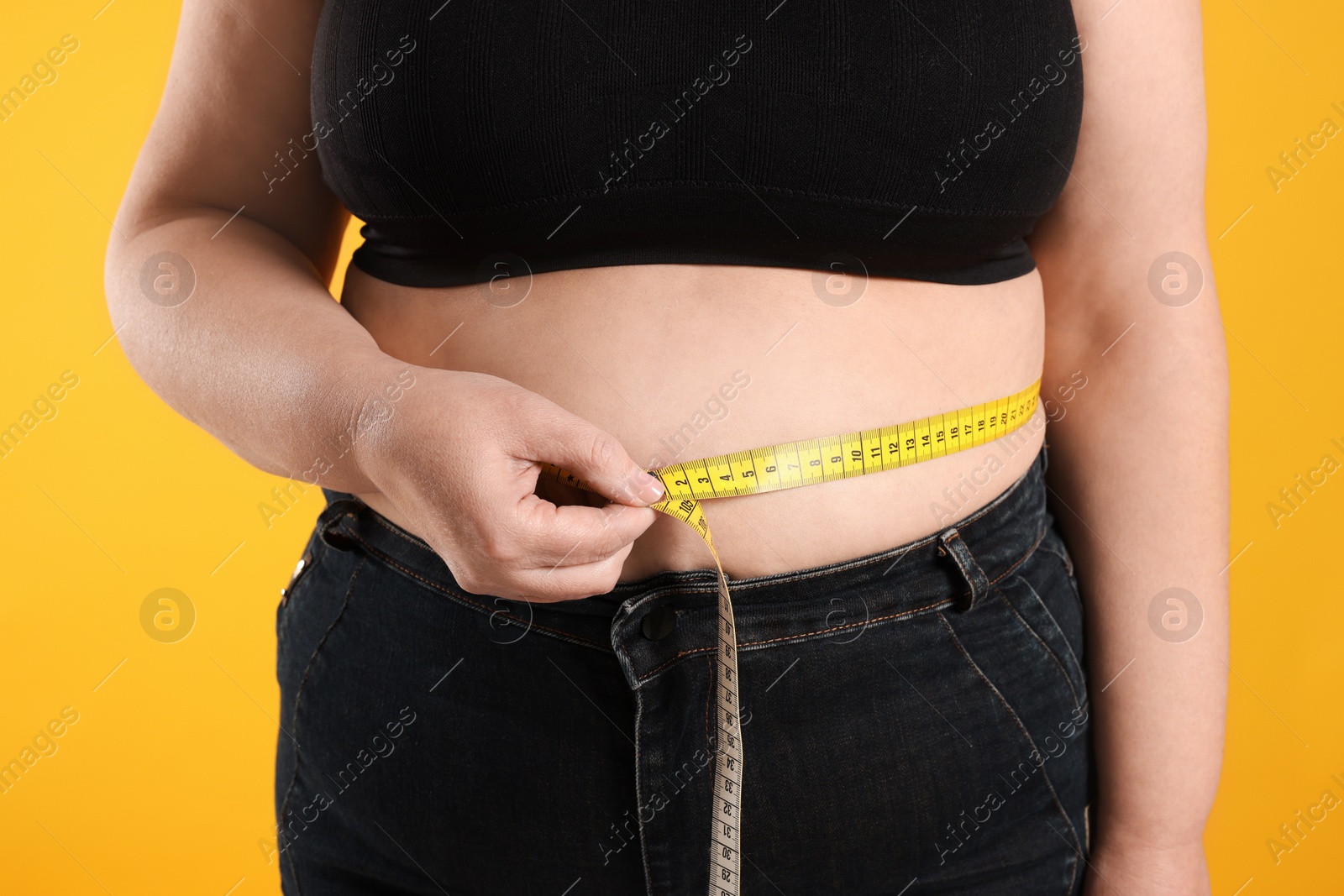 Photo of Overweight woman measuring waist with tape on orange background, closeup