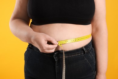 Photo of Overweight woman measuring waist with tape on orange background, closeup