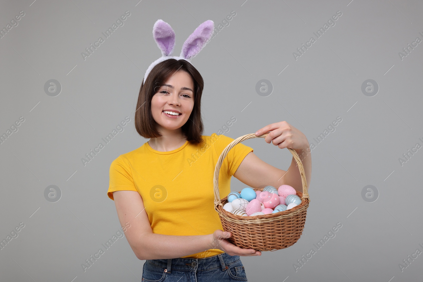 Photo of Easter celebration. Happy woman with bunny ears and wicker basket full of painted eggs on grey background