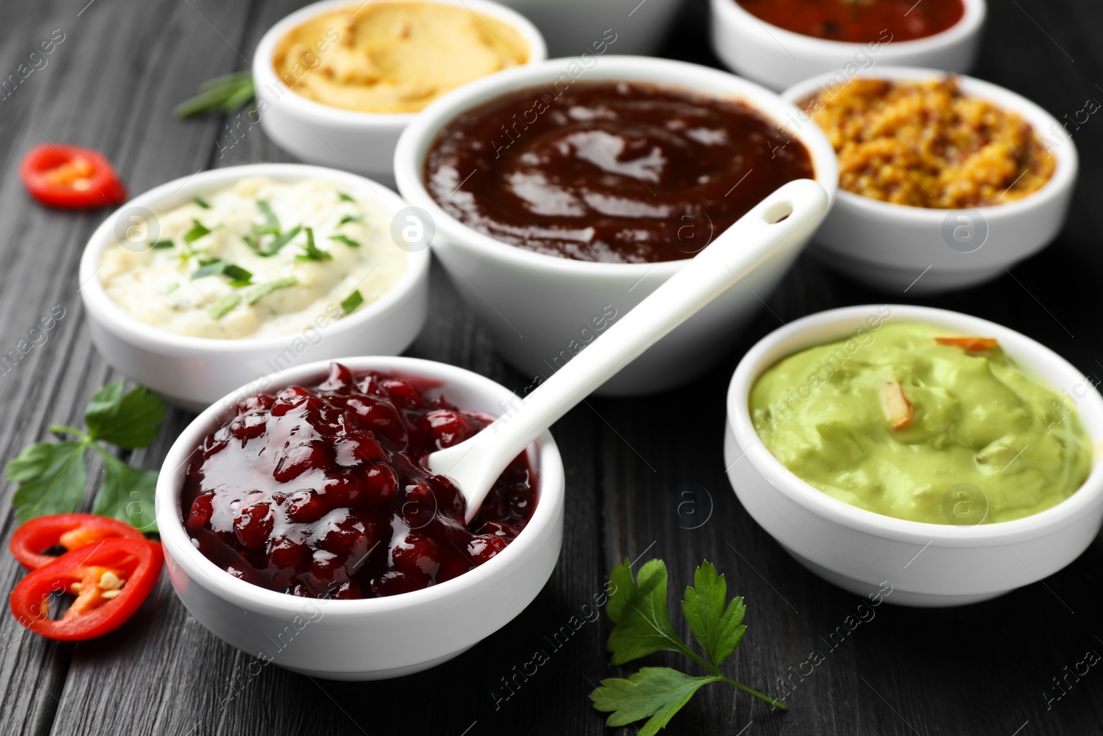 Photo of Different tasty sauces in bowls, parsley and chili pepper on black wooden table, closeup