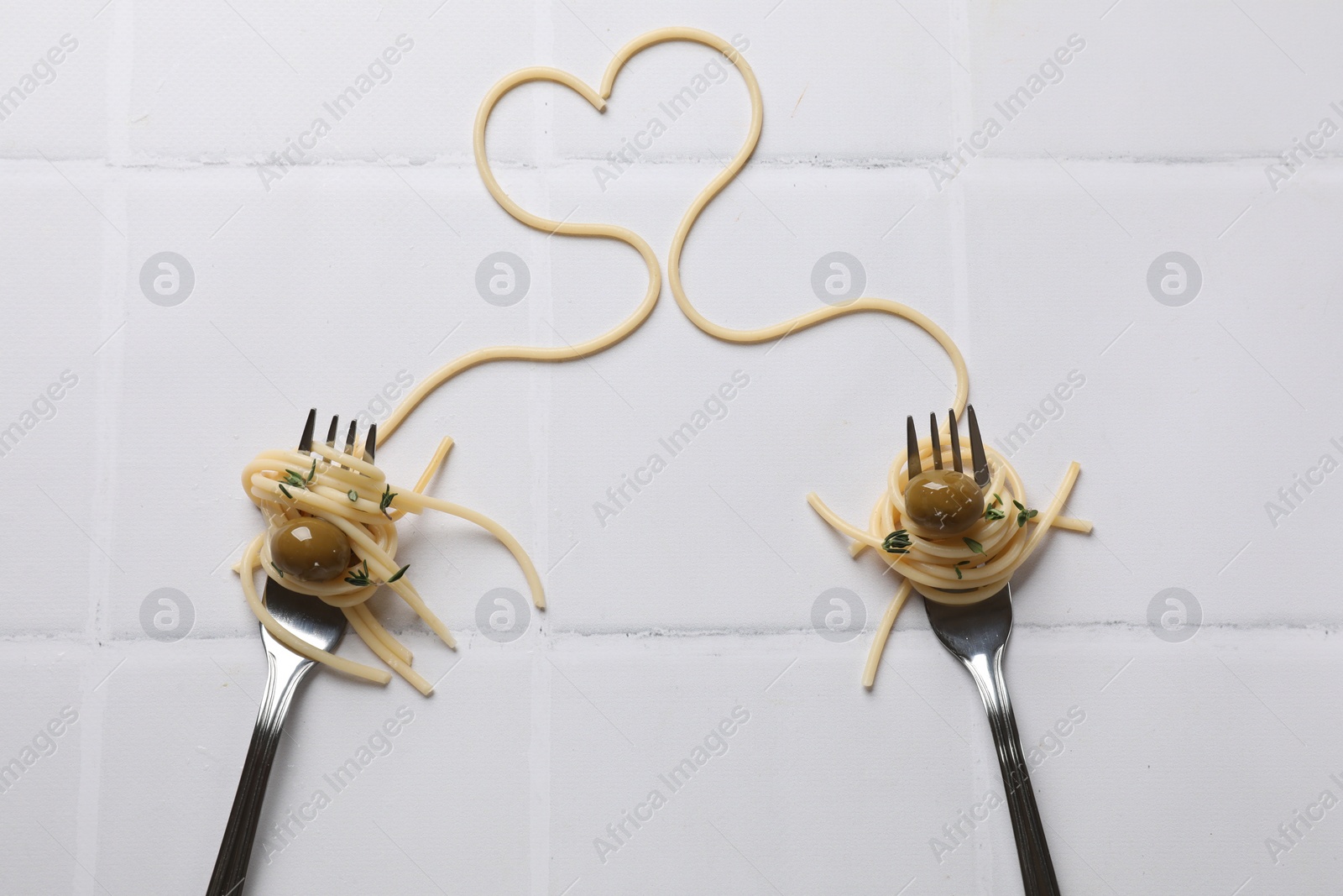 Photo of Heart made of tasty spaghetti, forks and olives on white tiled table, top view