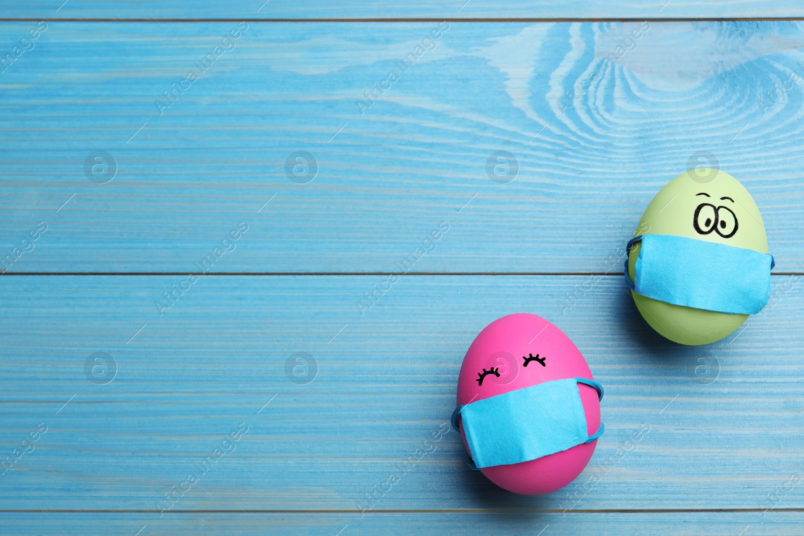 Photo of Bright eggs in protective masks on light blue wooden table, flat lay with space for text. Easter holiday during coronavirus quarantine