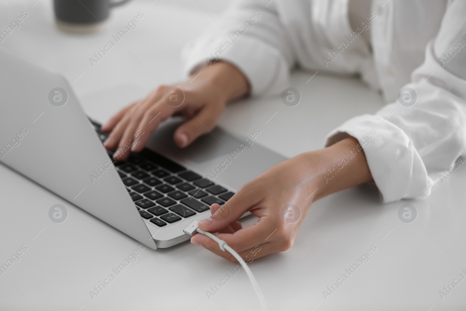 Photo of Woman connecting charger cable to laptop at white table, closeup