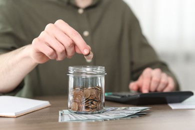 Financial savings. Man putting coin into glass jar while using calculator at wooden table, closeup