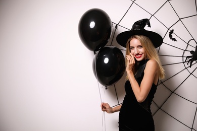 Woman in witch hat with balloons posing near white wall decorated for Halloween