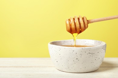 Photo of Pouring tasty honey from dipper into bowl at white wooden table against yellow background, closeup. Space for text