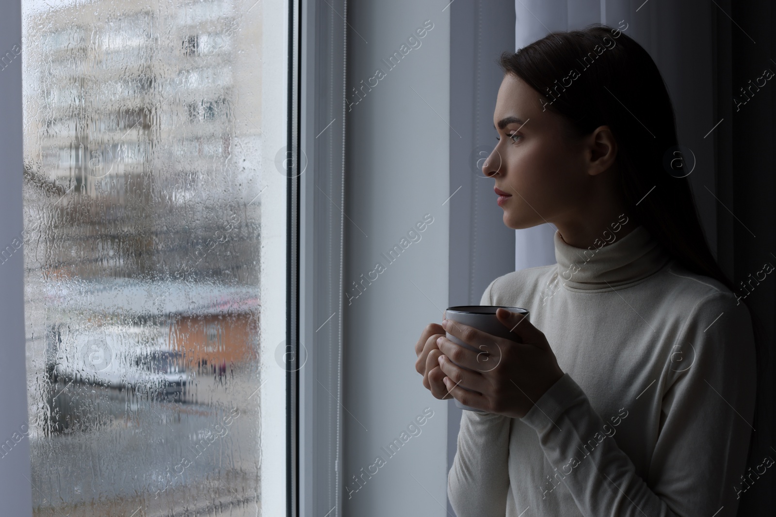 Photo of Melancholic young woman with drink looking out of window on rainy day, space for text. Loneliness concept