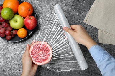 Photo of Woman putting plastic food wrap over half of grapefruit at grey table, top view