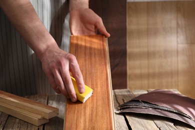 Photo of Man polishing wooden plank with sandpaper at table indoors, closeup