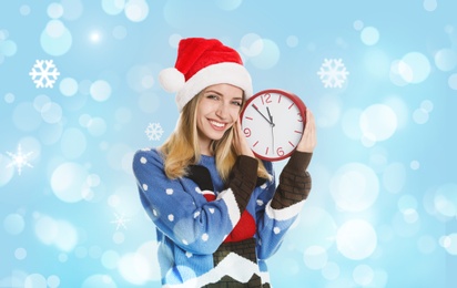 Image of New Year countdown. Happy woman in Santa hat holding clock on light blue background