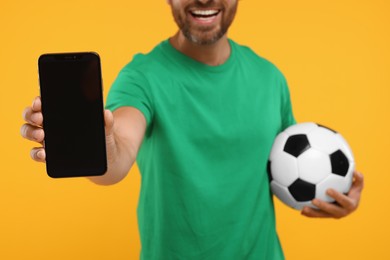 Photo of Happy sports fan with soccer ball and smartphone on orange background, closeup