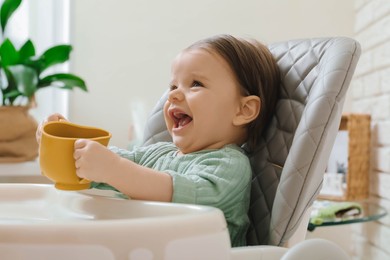 Photo of Cute little baby with cup in high chair indoors