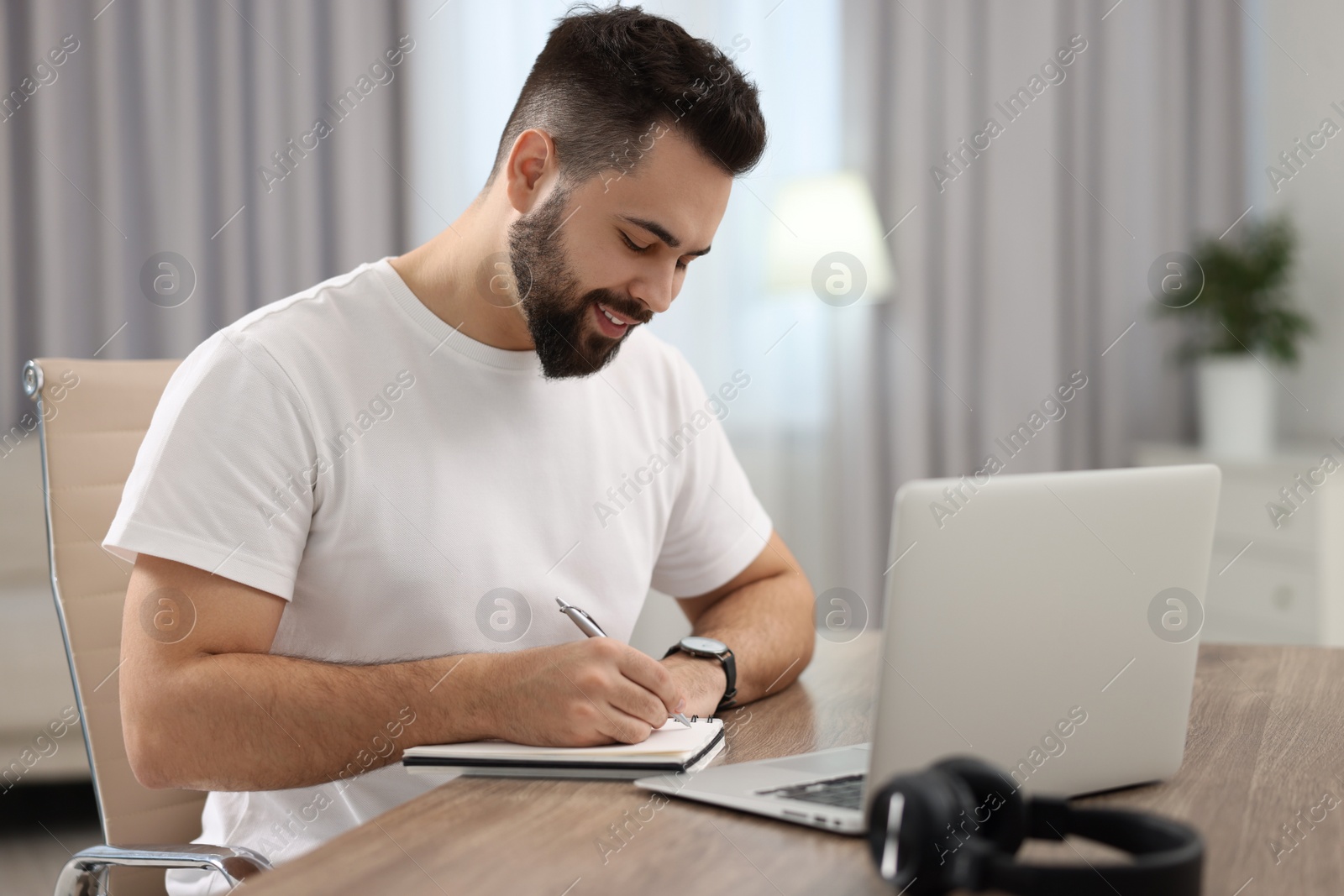 Photo of Young man writing down notes during webinar at table in room