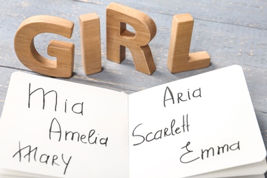 Notebook with written different baby names and wooden letters on grey wooden table, closeup