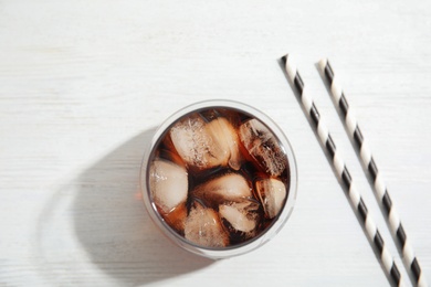Glass of refreshing cola with ice cubes on white wooden background, top view