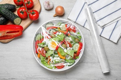 Photo of Bowl of fresh salad with plastic food wrap on white wooden table, flat lay