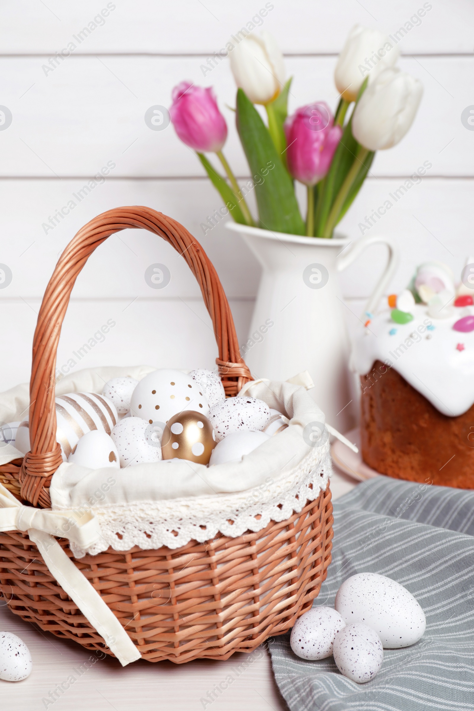 Photo of Wicker basket with festively decorated eggs, beautiful tulips and traditional Easter cake on white wooden table