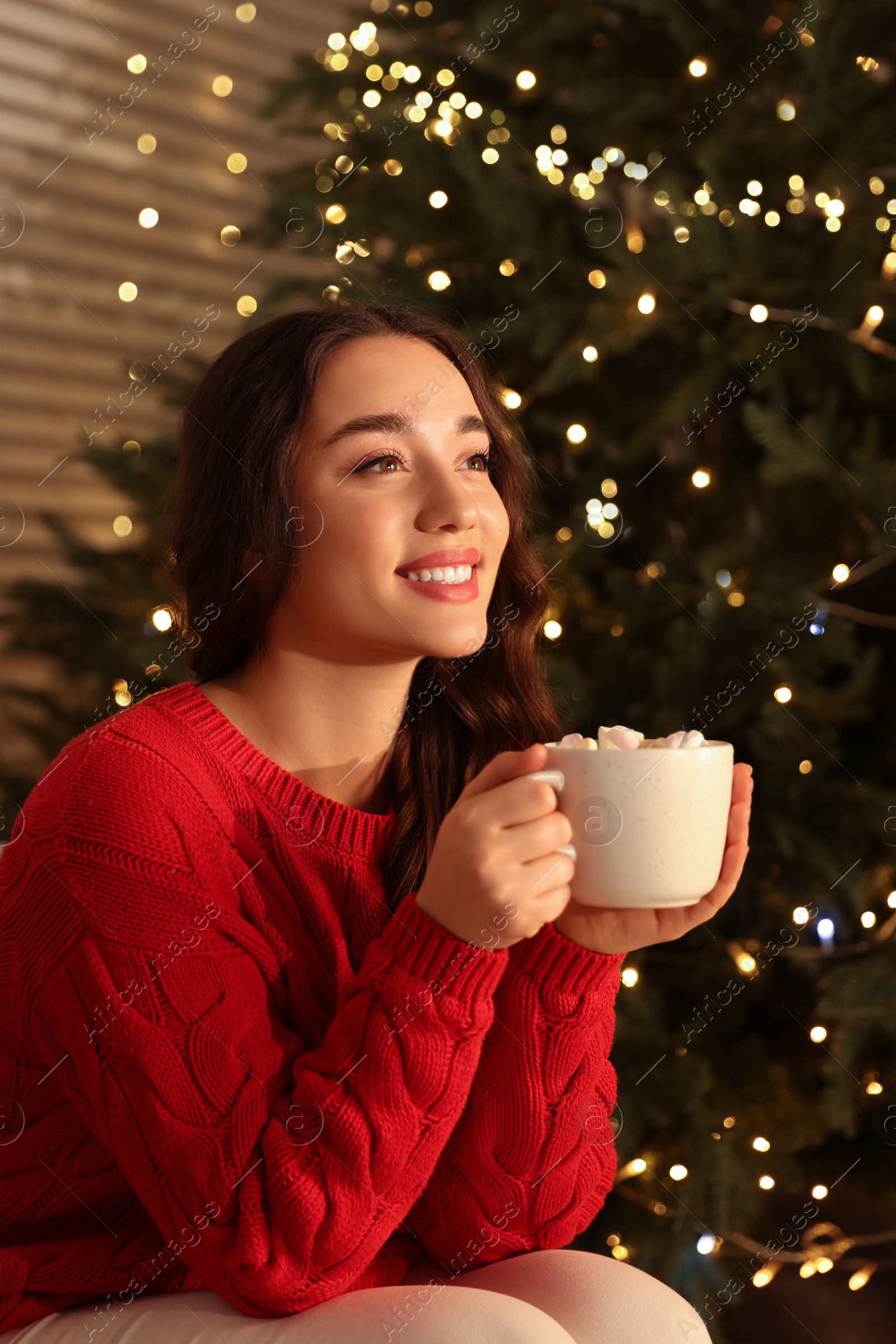 Photo of Beautiful woman with cup of tasty drink near Christmas tree indoors