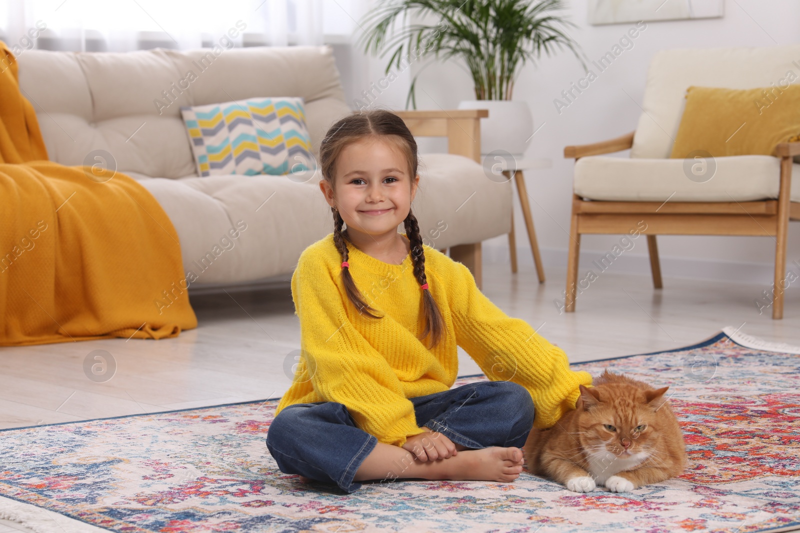 Photo of Smiling little girl petting cute ginger cat on carpet at home