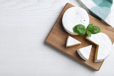 Photo of Tasty cut and whole brie cheeses with basil on white wooden table, flat lay. Space for text