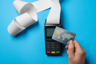 Photo of Man using payment terminal with credit card and thermal paper for receipt on light blue background, top view