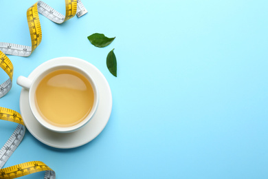 Photo of Cup of diet herbal tea, green leaves and measuring tape on light blue background, flat lay. Space for text