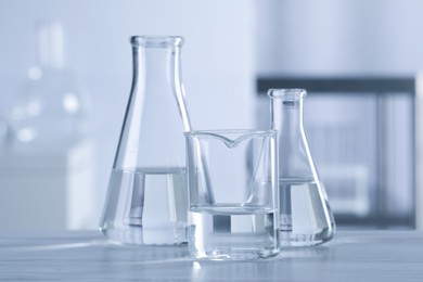Different laboratory glassware with transparent liquid on wooden table against blurred background
