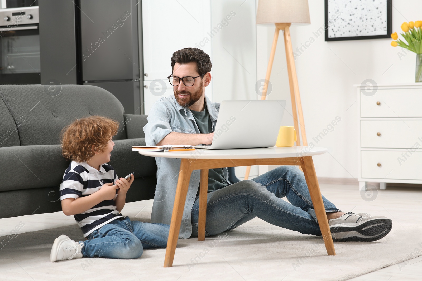 Photo of Man working remotely at home. Father using laptop at desk while his son playing with smartphone in living room