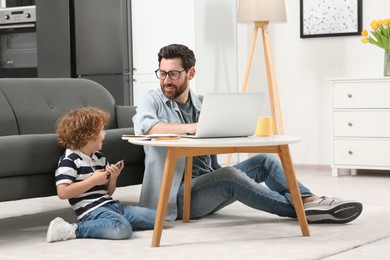 Photo of Man working remotely at home. Father using laptop at desk while his son playing with smartphone in living room