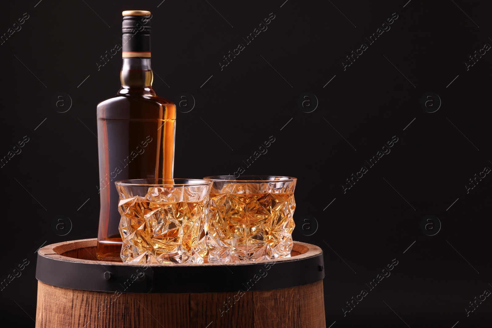 Photo of Whiskey in glasses and bottle on wooden barrel against black background, space for text