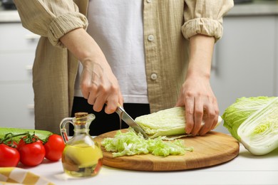 Woman cutting fresh chinese cabbage at white wooden table in kitchen, closeup