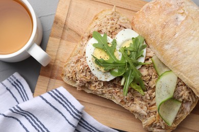 Delicious sandwich with tuna, boiled egg, vegetables and cup of tea on light grey table, flat lay