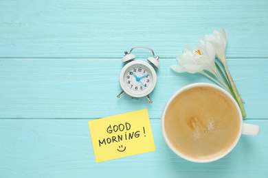 Photo of Delicious coffee, flowers and card with GOOD MORNING wish on light blue wooden table, flat lay. Space for text