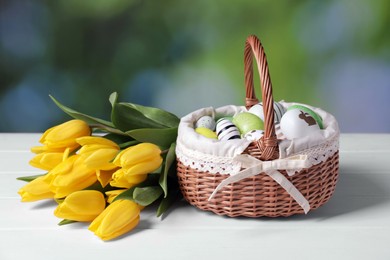 Photo of Wicker basket with festively decorated Easter eggs and beautiful tulips on white wooden table