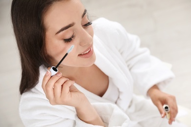 Photo of Attractive young woman applying oil onto her eyelashes indoors