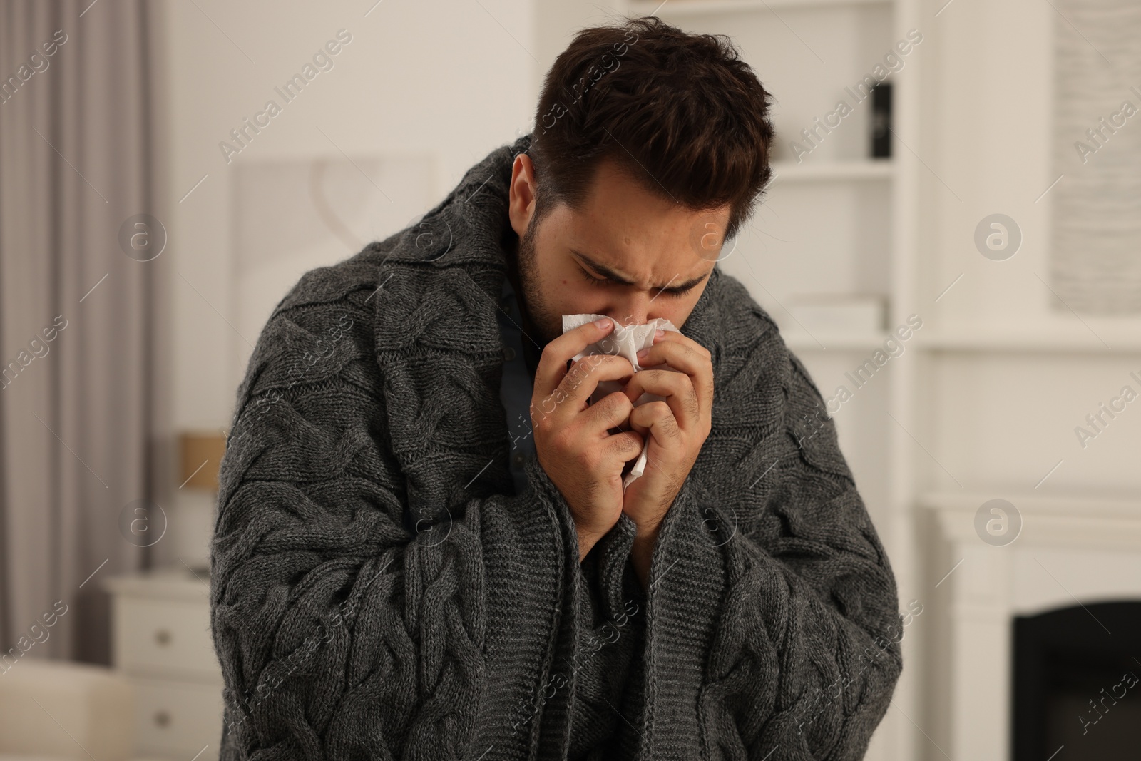 Photo of Sick man wrapped in blanket with tissue blowing nose at home. Cold symptoms