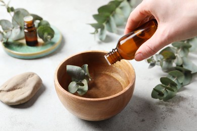 Photo of Woman dripping eucalyptus essential oil from bottle into bowl at light grey table, closeup