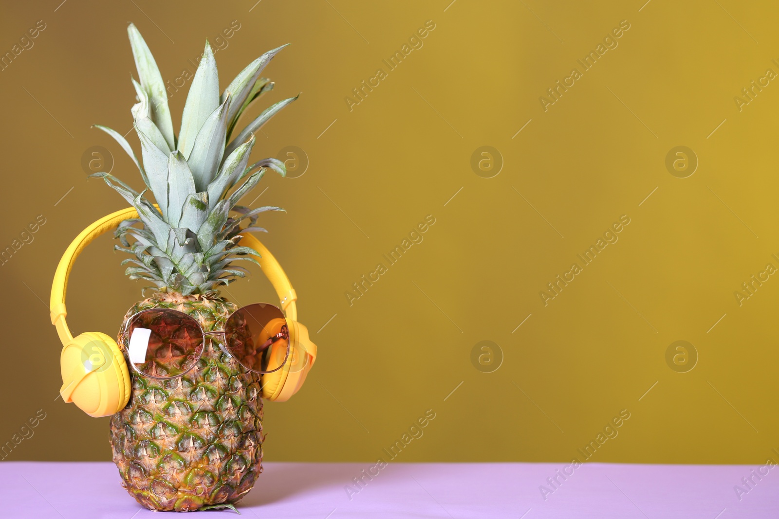 Photo of Pineapple with headphones and sunglasses on table against color background. Space for text