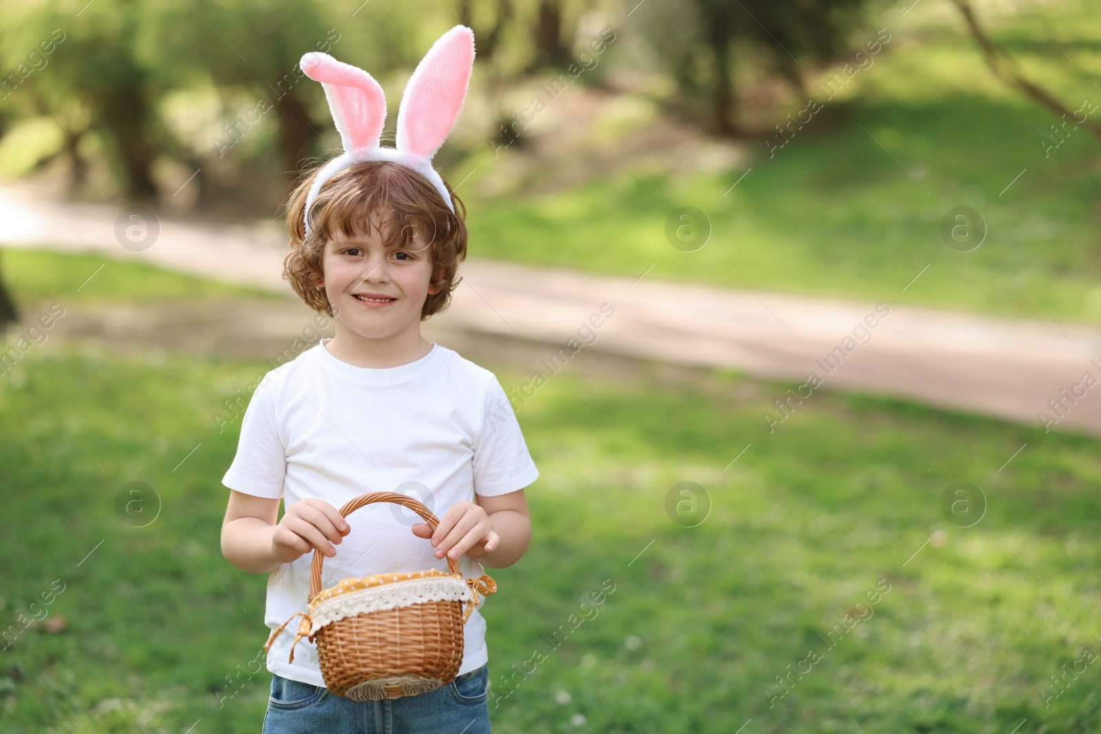 Photo of Easter celebration. Cute little boy in bunny ears holding wicker basket outdoors, space for text