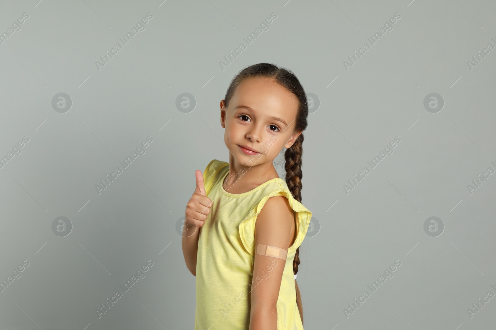 Photo of Vaccinated little girl with medical plaster on her arm showing thumb up against light grey background