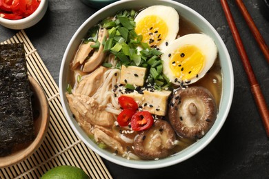 Bowl of delicious ramen, ingredients and chopsticks on black table, flat lay. Noodle soup