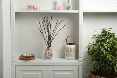 Beautiful pussy willow branches in vase with painted eggs on white shelf indoors. Easter decor