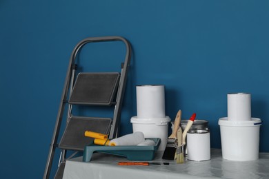 Photo of Metallic folding ladder and painting tools on table near blue wall