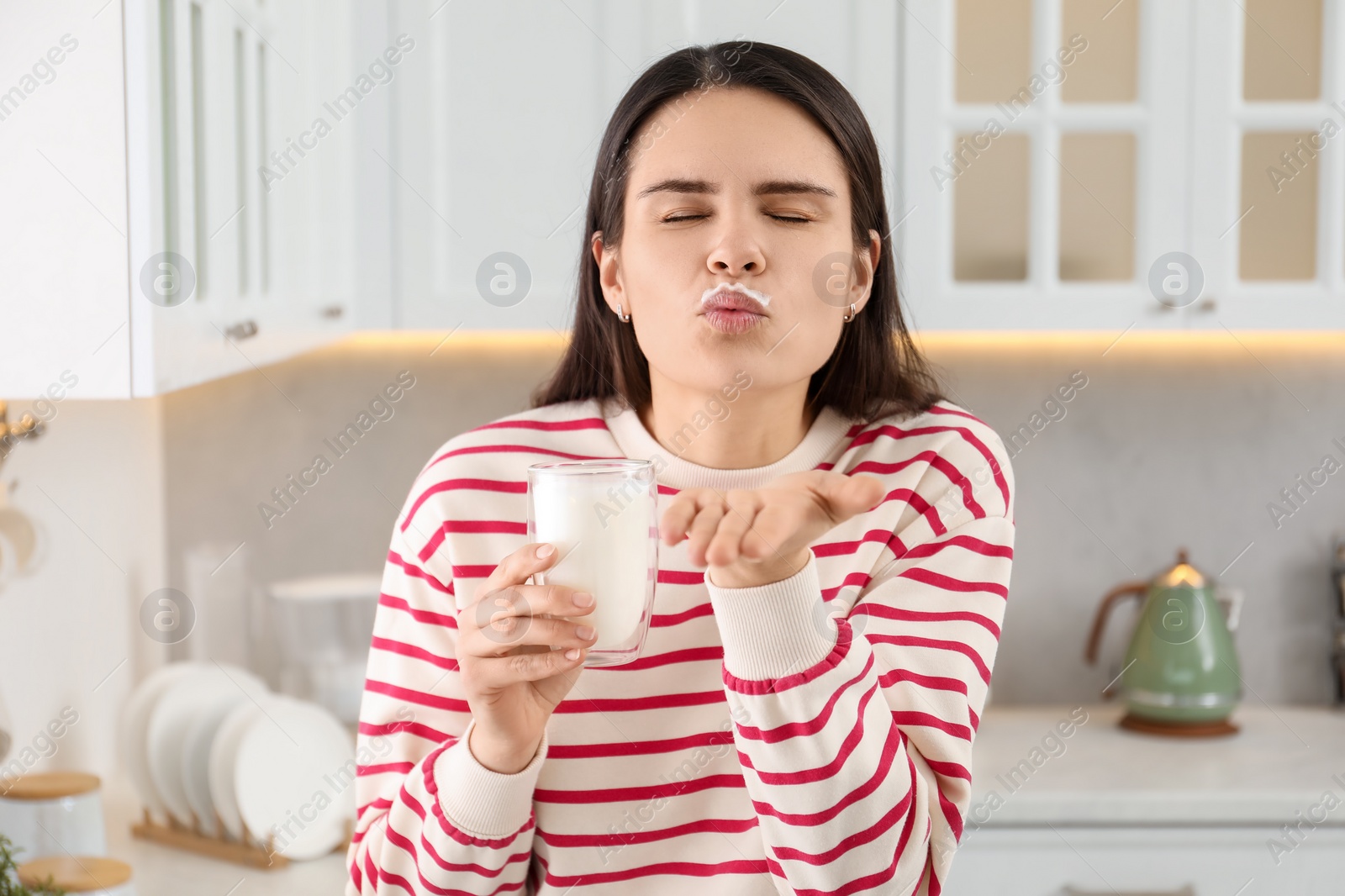 Photo of Happy woman with milk mustache holding glass of tasty dairy drink and sending air kiss in kitchen