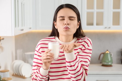 Happy woman with milk mustache holding glass of tasty dairy drink and sending air kiss in kitchen